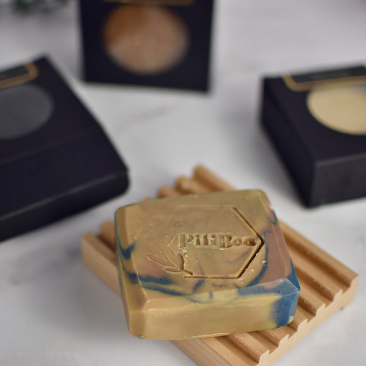 Bar of soap with a brand logo on a wooden soap dish, with blurred boxes in the background.