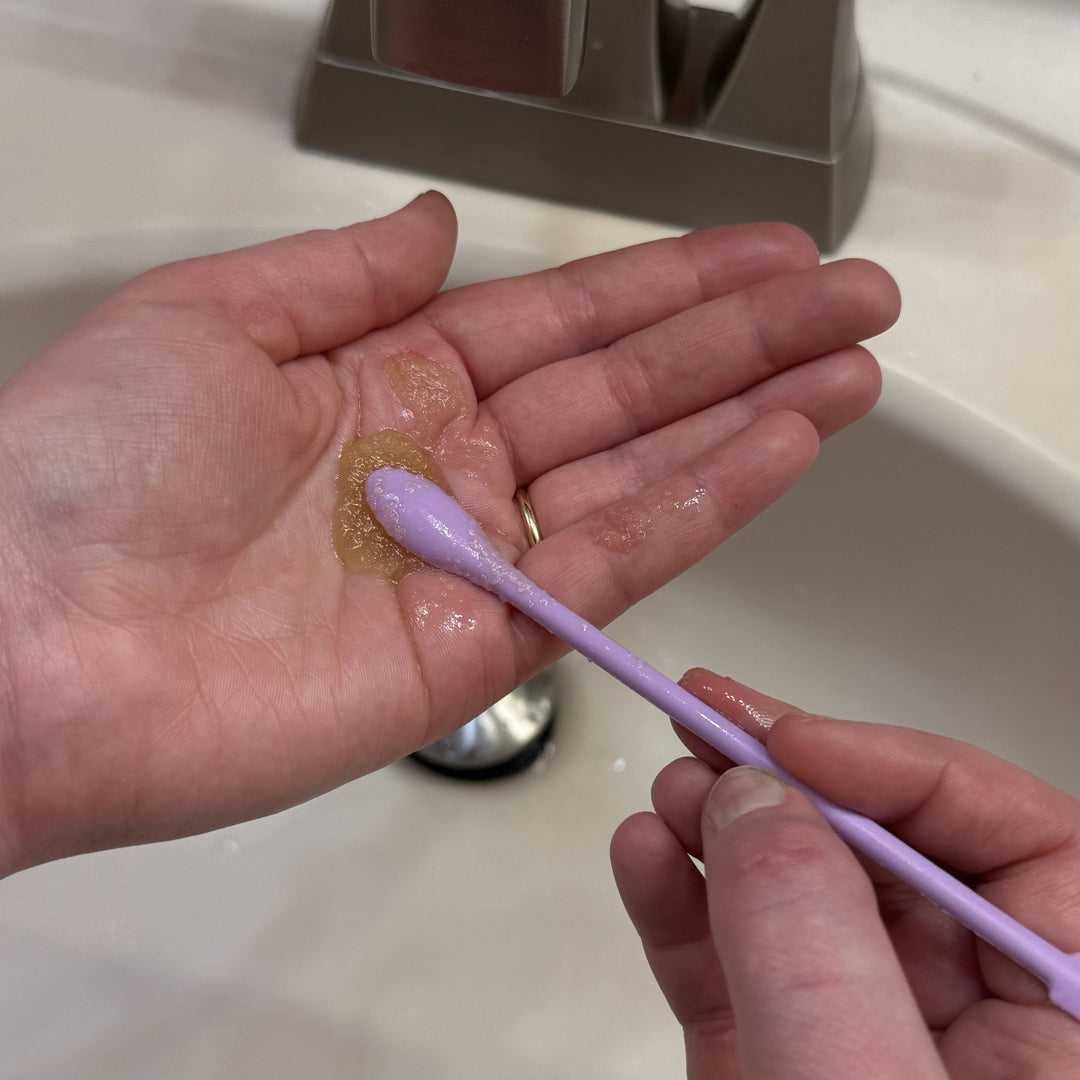 Person holding a small amount of gold balm on a cotton swab over their palm with a sink in the background.